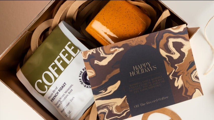 praznicna darila-3000x1134 2-1200x454 Na sliki je rjava škatla. V njej je zeleno bela vrečka kave in oranžna skodelica. Zraven je še rjavo črna voščilnica s posvetilom.