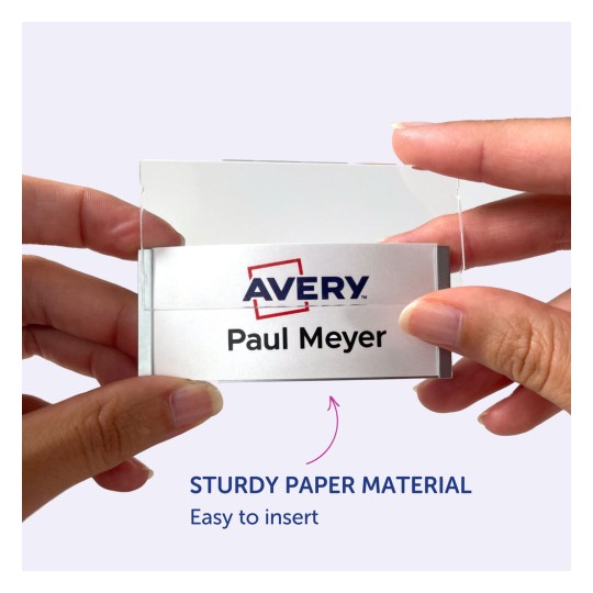 Slika prikazuje osebo, ki drži priponko z imenom in logotipom Avery, na kateri je napisano 'Paul Meyer'. Zdi se, da je priponko enostavno vstaviti v držalo iz trpežnega papirnatega materiala.