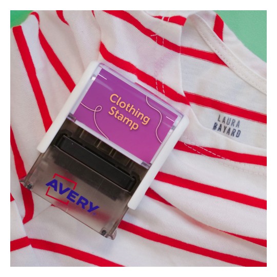 Slika prikazuje oblačilni žig z napisom 'Clothing Stamp', nameščen na majico s črtami, ki ima rdeče črte. Majica ima oznako z napisom 'LAURA BAYARD'. Žig je znamke 'Avery'.