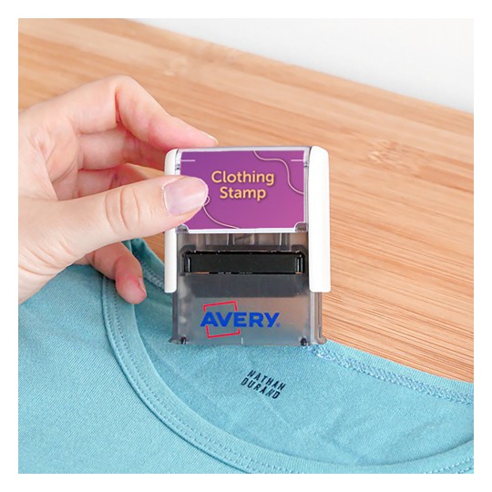 Slika prikazuje osebo, ki drži žig za oblačila, postavljen nad majico. Žig je označen z 'Clothing Stamp' in ima vidno blagovno znamko 'Avery'.