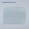 Slika prikazuje jasno, prozorno plastično držalo za kartice z zaobljenimi robovi in luknjami za pritrditev na vrhu. Nima nobene vstavljene vsebine in je postavljeno na sivo ozadje.
