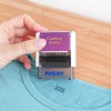 Slika prikazuje osebo, ki drži žig za oblačila, postavljen nad majico. Žig je označen z 'Clothing Stamp' in ima vidno blagovno znamko 'Avery'.